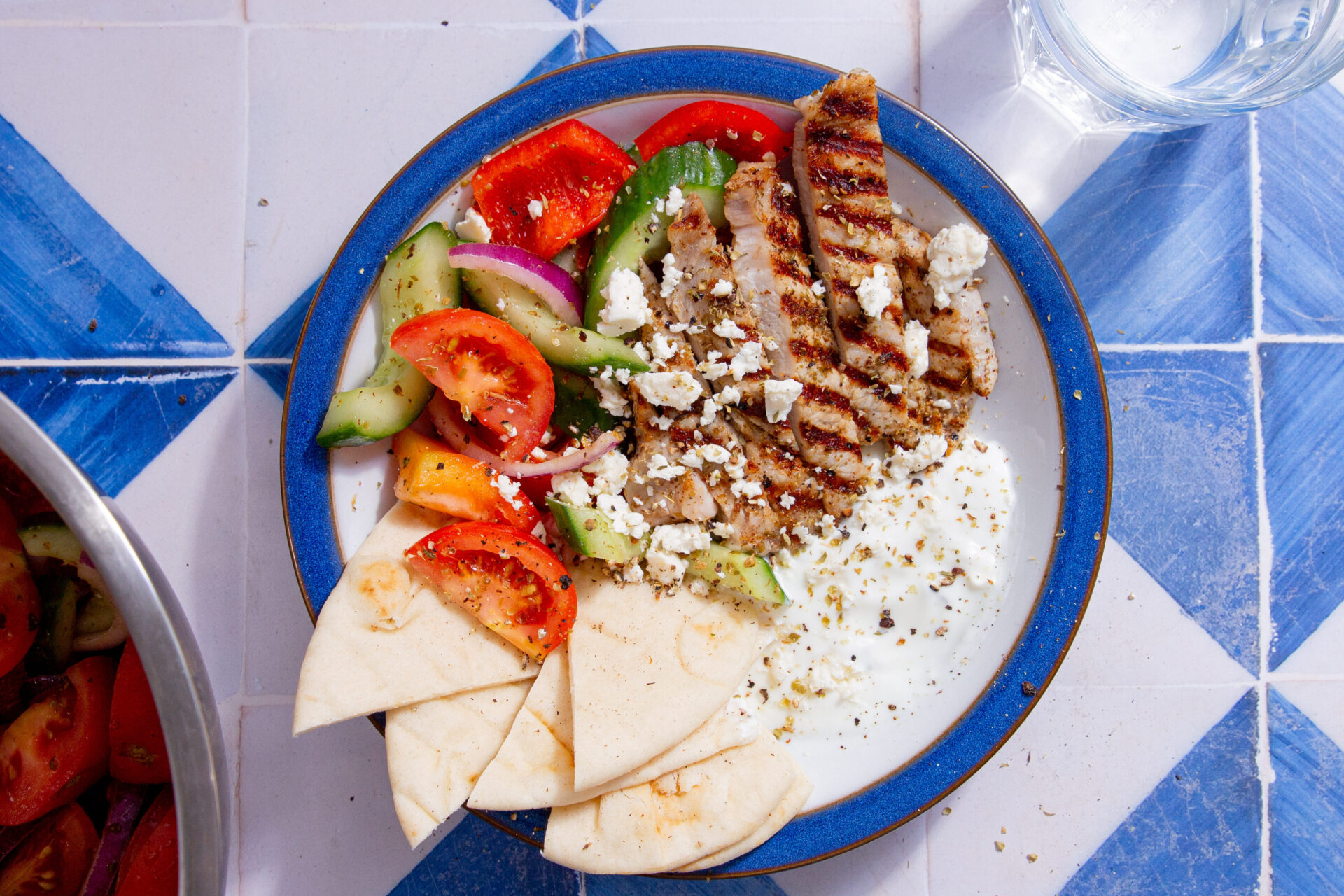 Pork Gyros bowl with pork, pitta bread, salad and dip in a bowl on a blue and white tiled background