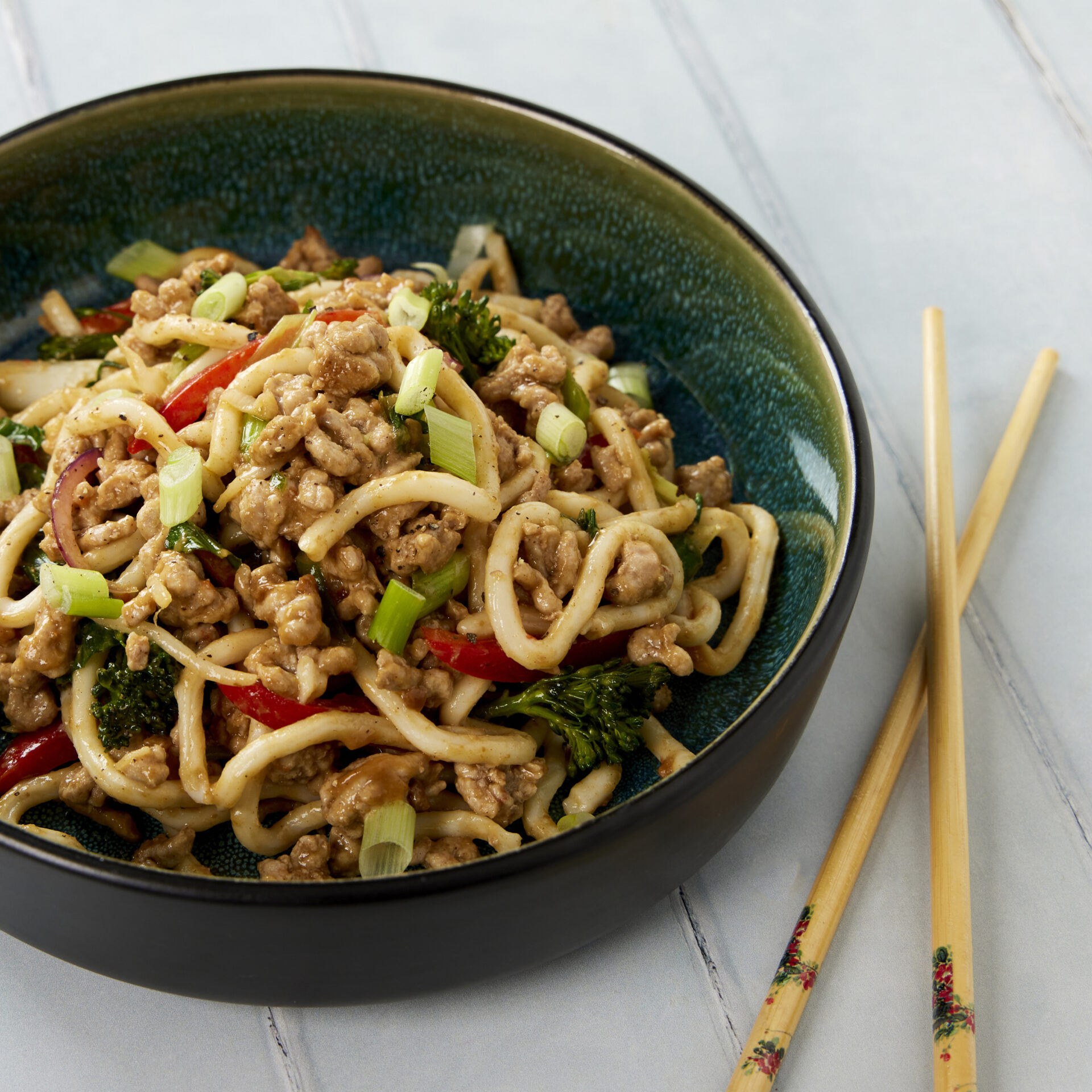 A close up shot of a bowl of udon noodles, with pork mince and vegetables. The bowl is next to a set of chopsticks.