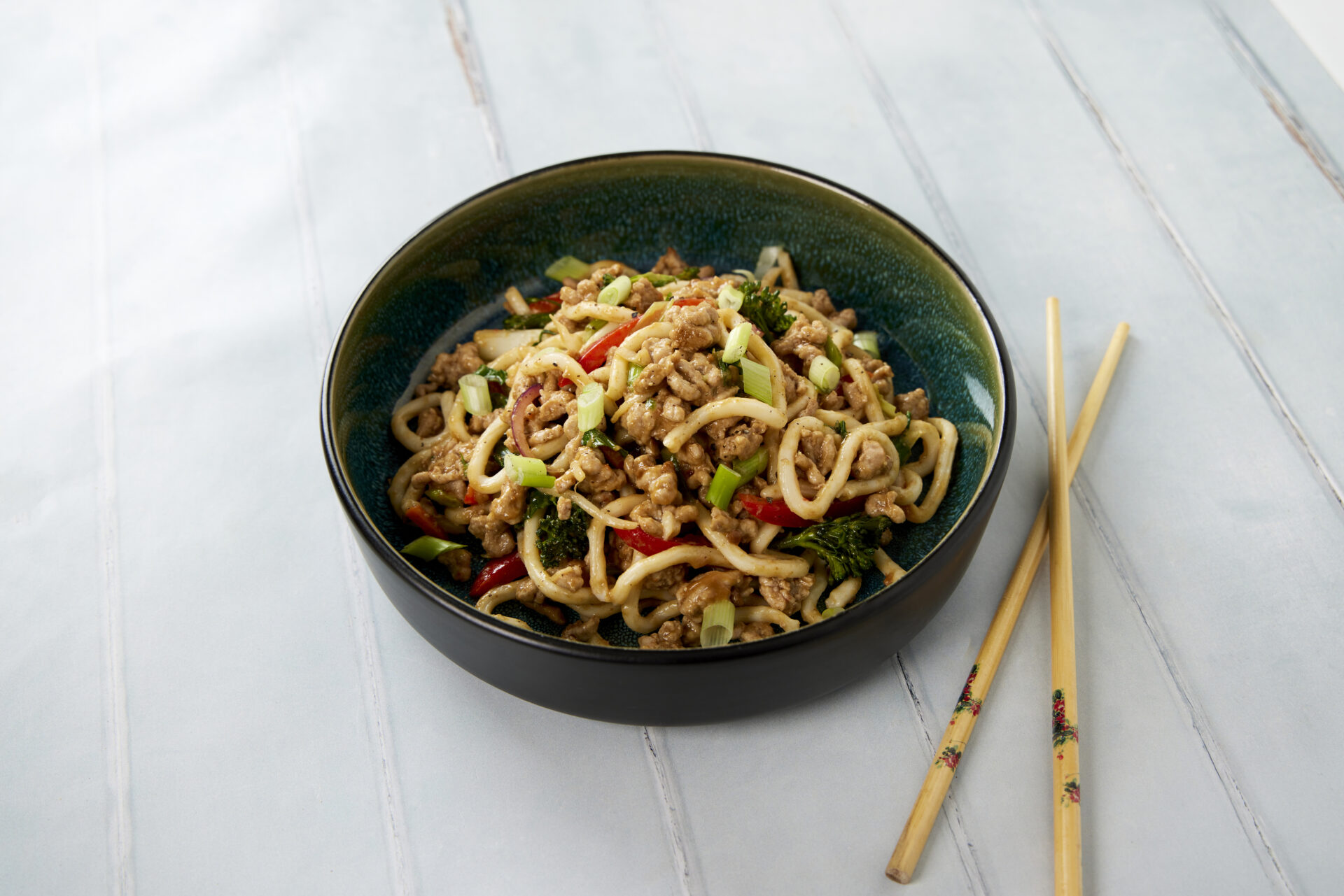 A bowl of udon noodles, with pork mince and vegetables. The bowl is next to a set of chopsticks.