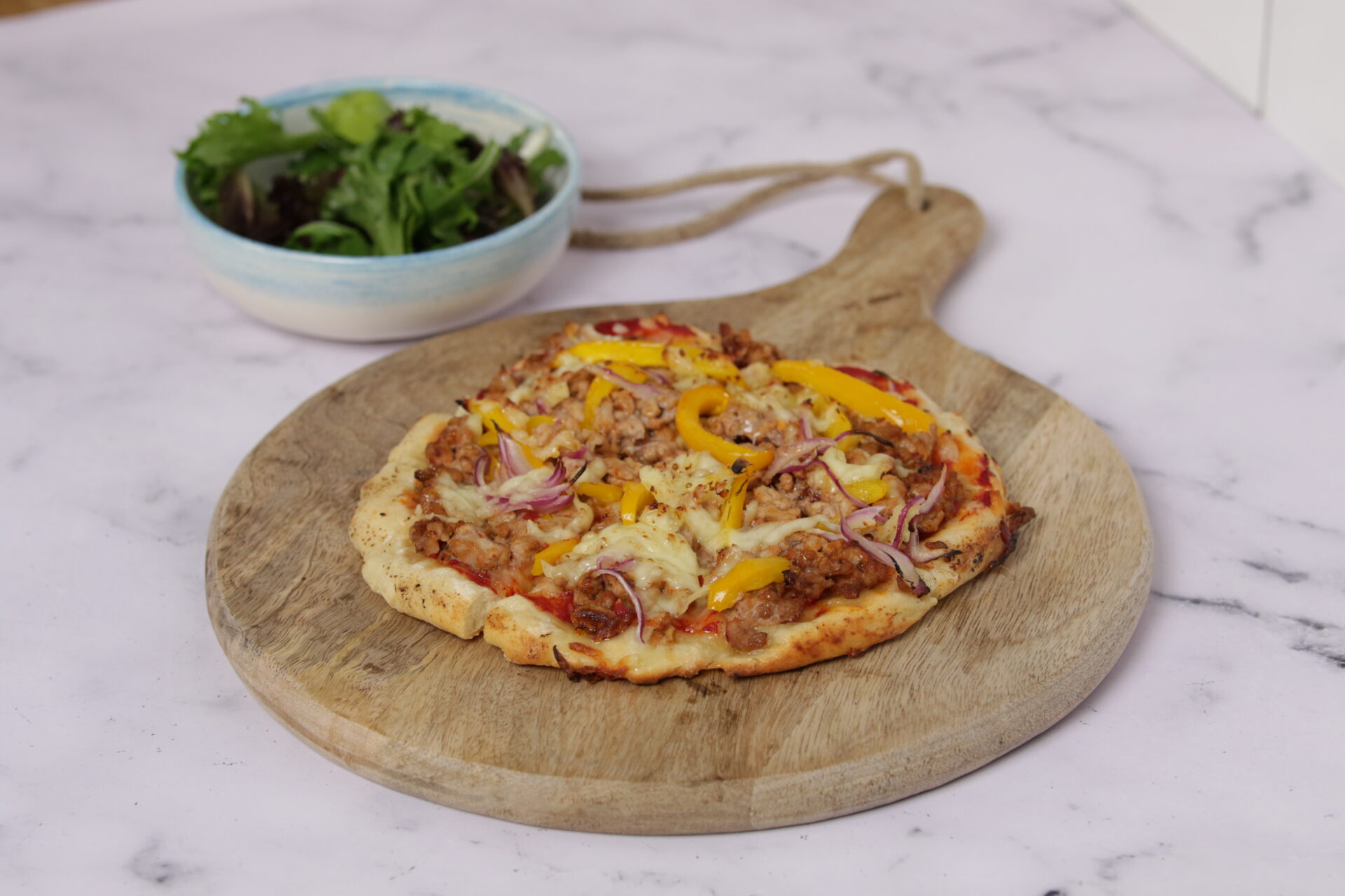 A wooden board holding a pizza, which is topped with pork mince, and greens on the side.